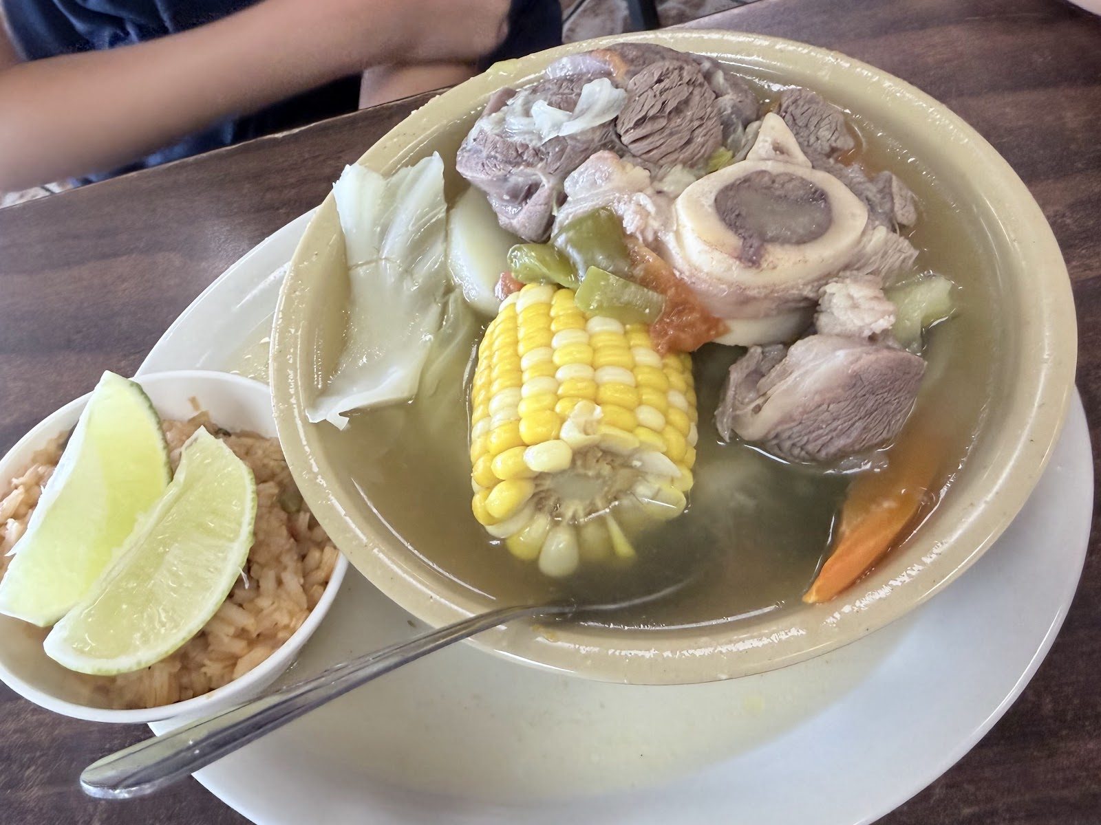 Bowl of caldo de res with a beef shank on the bone, corn on the cob and vegetables, with rice and lime