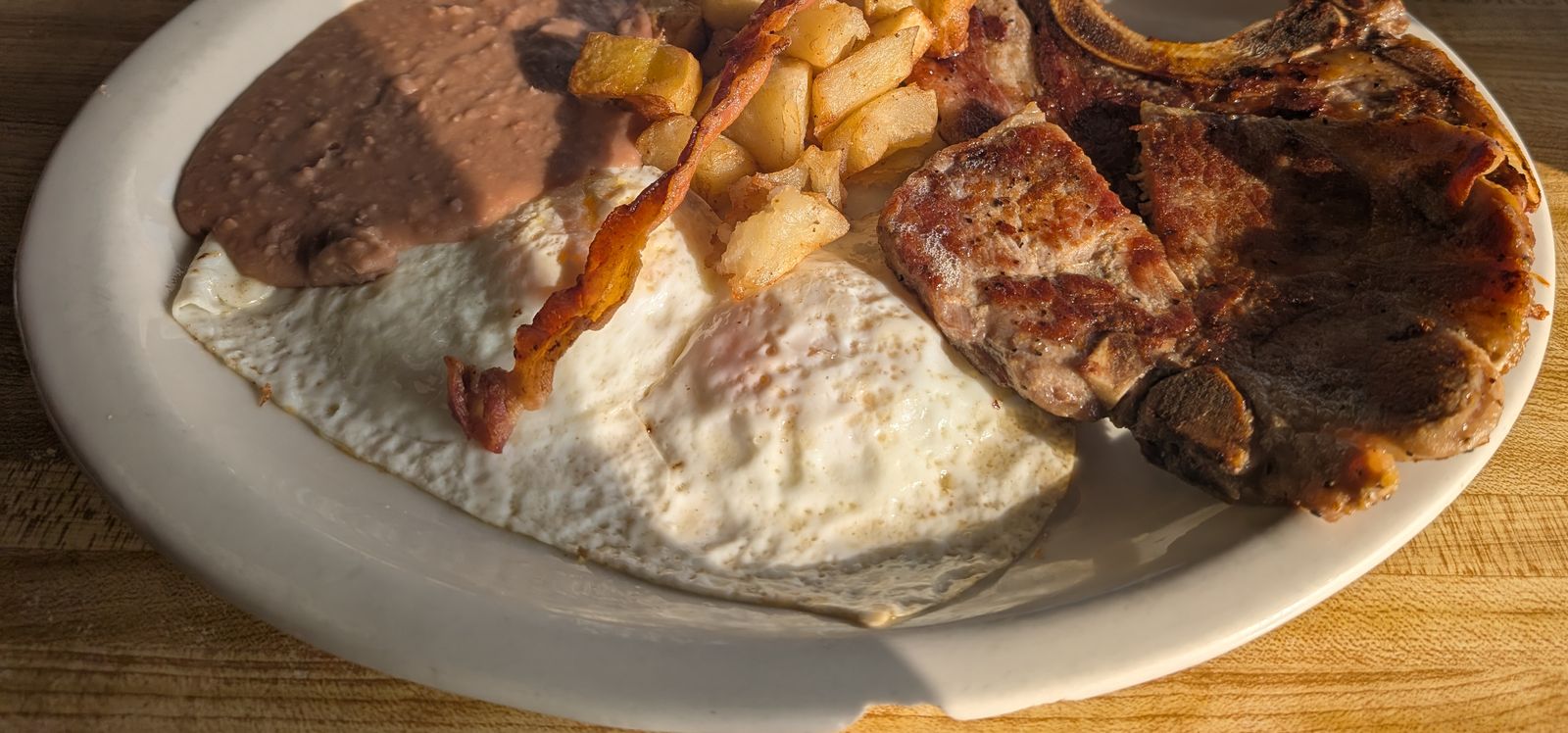 Plate with a bone-in pork chop, fried eggs, bacon, fried potatoes and refried beans